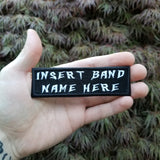 White on black rectangular embroidered patch that says insert band name here, shown in the palm of a hand for size reference.