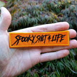Orange patch with the words spooky shit for life embroidered in black thread.