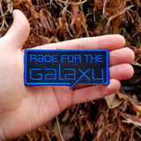 Blue on black embroidered logo patch for the classic board game Race for the Galaxy, shown in the palm of a hand for size reference.