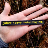 An embroidered patch reading slow heavy metal playing, based on the classic internet meme of a movie subtitle, shown in the palm of a hand for size reference.