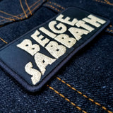 Close up detail of a rectangular tan on black embroidered patch with the words beige sabbath in the logo style of the classic rock band black sabbath.