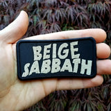 Rectangular tan on black embroidered patch with the words beige sabbath in the logo style of the classic rock band black sabbath. Shown in the palm of a hand for size reference.