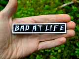 Rectangular white on black embroidered patch with the phrase bad at life. Shown in the palm of a hand for size reference.
