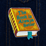 Embroidered patch of a hardcover book with a brown cover and gold detail, the front title says the book is always better in green thread. Shown on a denim jacket.