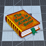 Embroidered patch of a hardcover book with a brown cover and gold detail, the front title says the book is always better in green thread.