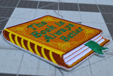 Close up detail of an embroidered patch of a hardcover book with a brown cover and gold detail, the front title says the book is always better in green thread.