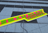 Close up detail of a narrow, sharp angled embroidered patch that says sexually mediocre in bright neon green, pink, and yellow colors.