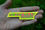 A narrow, sharp angled embroidered patch that says sexually mediocre in bright neon green, pink, and yellow colors. Shown in the palm of a hand for size reference.