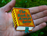 Embroidered patch of a hardcover book with a brown cover and gold detail, the front title says the book is always better in green thread. Shown in the palm of a hand for size reference.