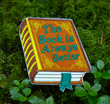 Embroidered patch of a hardcover book with a brown cover and gold detail, the front title says the book is always better in green thread.