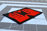 Close up detail of a rectangular black embroidered patch of the red hands of Manos, the Hands of Fate.