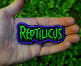 Embroidered patch with 'REPTILICUS' text in green on a black background with purple border. Shown in the palm of a hand for size reference.