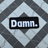 Rectangular black patch with white embroidery spelling the word damn with a period at the end.