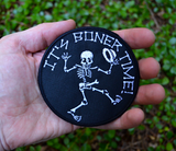 Round black embroidered patch with a dancing skeleton and the words it's boner time in white thread. Shown in the palm of a hand.