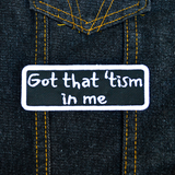 Rectangular black patch with a white border and the words, got that tism in me, embroidered in white thread. Shown on a denim jacket.