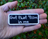 Rectangular black patch with a white border and the words, got that tism in me, embroidered in white thread. Shown in the palm of a hand for size reference.