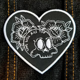 Heart shaped white on black embroidered patch with two flowers and a skull with the words love will kill you written on it.