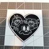 Heart shaped white on black embroidered patch with two flowers and a skull with the words love will kill you written on it.