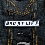 Bad At Life embroidered patch in black on white.