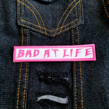 Bad At Life embroidered patch in pink on white.