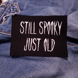 White on black screen printed canvas patch that says still spooky, just old.