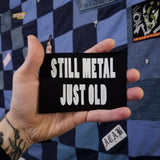 White on black screen printed canvas patch that says still metal, just old. Shown in the palm of a hand for size reference.