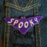 Purple bat-shaped felt patch with the word spooky sewn in multicolor thread.
