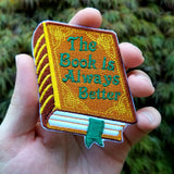 Embroidered book that says, the book is always better, on the cover, held in a hand for size reference.