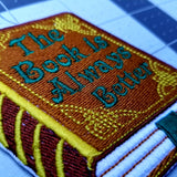 Close up detail of an embroidered book that says, the book is always better, on the cover.