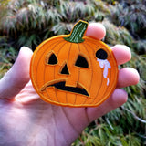 An orange embroidered patch of a sad-looking jack o lantern with a hole in the side of it and white liquid dripping out.