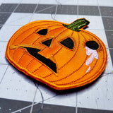 An orange embroidered patch of a sad-looking jack o lantern with a hole in the side of it and white liquid dripping out.