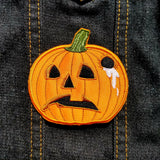 An orange embroidered patch of a sad-looking jack o lantern with a hole in the side of it and white liquid dripping out.