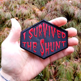 Red on black embroidered patch that says I survived the shunt, in reference to the horror movie Society, shown in the palm of a hand for size reference.