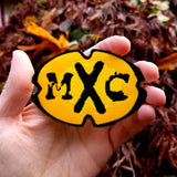 The MXC (Most Extreme Elimination) logo as an embroidered patch. Shown in a hand for size reference.