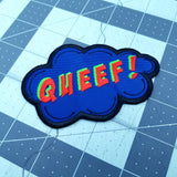 An embroidered patch in the shape of a comic book sound effect bubble with the word queef.