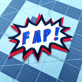 An embroidered patch in the shape of a comic book sound effect bubble with the word fap.