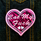 A heart-shaped pink and white embroidered patch on zebra print fabric with the phrase Eat My Fuck.