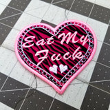 A heart-shaped embroidered Valentines patch with black and pink leopard print, white lace border, and the words eat my fuck.