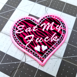 A heart-shaped pink and white embroidered patch on zebra print fabric with the phrase Eat My Fuck.