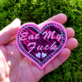 A heart-shaped embroidered Valentines patch with black and pink leopard print, white lace border, and the words eat my fuck. Shown in the palm of a hand for size reference.