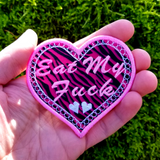 A heart-shaped pink and white embroidered patch on zebra print fabric with the phrase Eat My Fuck. Shown in the palm of a hand for size reference.
