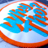 Close up detail of a circular embroidered patch with glittery blue and flat orange lettering saying why though on white fabric.