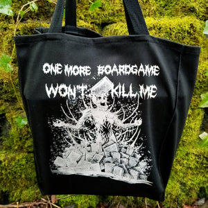 A black canvas tote bag with a white screen printed graphic of a zombie in a pile of boardgames and a caption saying one more boardgame won't kill me.