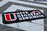Close up detail of a rectangular black embroidered patch with the words unite, unionize, undermine written in a stencil font, featuring a red sickle.