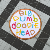 Circular embroidered patch with the words big dumb doodie head in multiple colors written with a childish font.