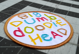 Close up detail of a circular embroidered patch with the words big dumb doodie head in multiple colors written with a childish font.