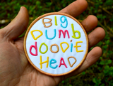 Circular embroidered patch with the words big dumb doodie head in multiple colors written with a childish font. Shown in the palm of a hand for size reference.