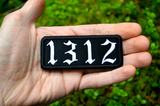 A white on black rectangular embroidered patches with the number 1312. Shown in the palm of a hand for size reference.