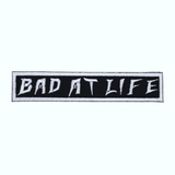 Rectangular white on black embroidered patch with the phrase bad at life.