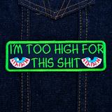 Rectangular green-bordered embroidered patch with a pair of bloodshot eyes and the text, I'm too high for this shit in green thread. Shown on a denim jacket.